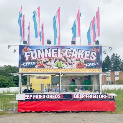 A colorful food stand featuring funnel cakes and deep-fried Oreos, with flags and two staff members visible.