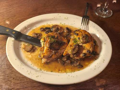 A plate of chicken breast topped with mushrooms in a savory sauce, garnished with herbs, and accompanied by a knife and fork.