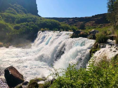 A cascading waterfall surrounded by lush greenery and rocky terrain under a clear blue sky.