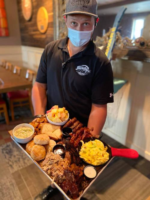 A server in a mask holds a large platter of assorted meats, sides, and fruits in a restaurant setting.