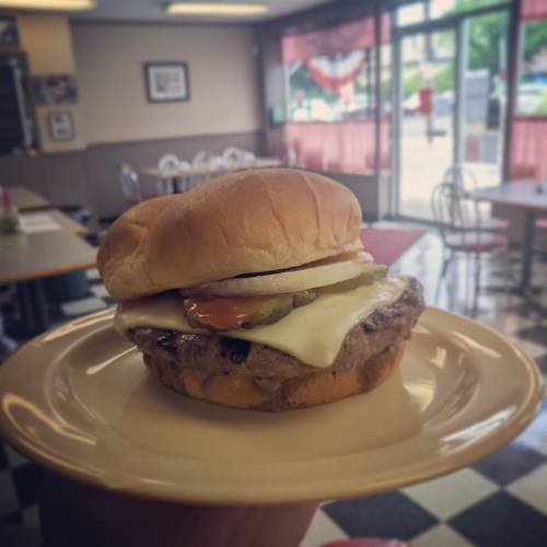 A close-up of a juicy burger with cheese, pickles, and onions on a bun, held on a plate in a diner setting.