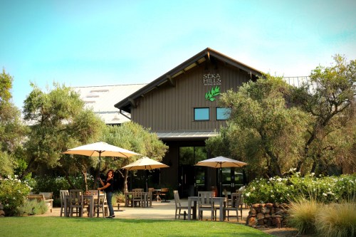 A woman stands outside Seka Hills, a rustic building with outdoor seating and umbrellas, surrounded by greenery.