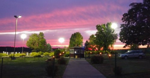 A vibrant sunset with purple and pink hues over a park, featuring lights, trees, and a pathway leading to a building.