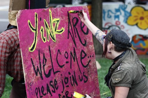 A person paints a bright pink sign that reads "Yinz Are Welcome... Please Join Us" at an outdoor event.