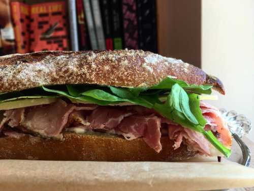 A close-up of a sandwich with layers of ham, spinach, and bread, set against a backdrop of books.