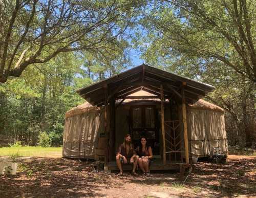 A couple sits on the steps of a yurt surrounded by trees on a sunny day.