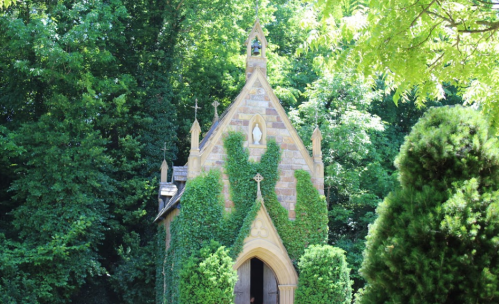 A small, ivy-covered chapel surrounded by lush green trees on a sunny day.