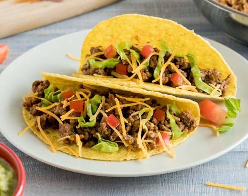 Two tacos filled with seasoned ground beef, lettuce, diced tomatoes, and shredded cheese on a white plate.