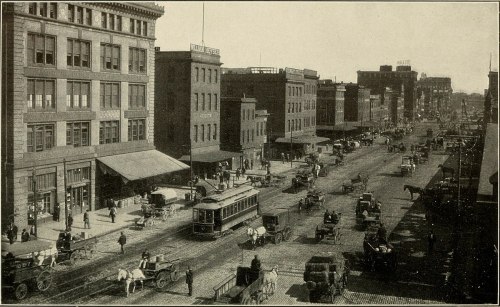 Historic street scene featuring horse-drawn carriages, a tram, and early 20th-century buildings.