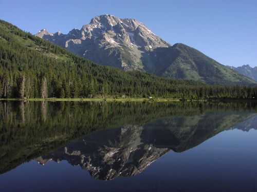 A serene mountain landscape with a clear lake reflecting the peaks and lush green trees under a blue sky.