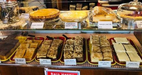 A display case filled with various desserts, including cakes and bars, labeled with names and prices.