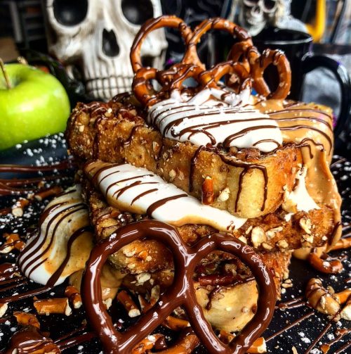 A decadent dessert featuring stacked bread topped with chocolate, caramel, pretzels, and drizzled sauces, with a skull in the background.