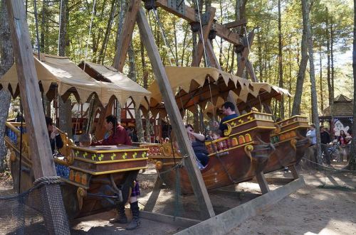 Two pirate ship rides swing back and forth in a wooded amusement park, with people enjoying the ride.