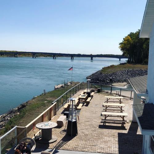 A scenic view of a river with a bridge in the background, featuring outdoor seating and an American flag.