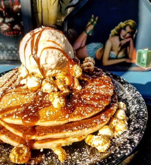Stack of pancakes topped with ice cream, caramel drizzle, and popcorn, with a vintage pin-up girl in the background.