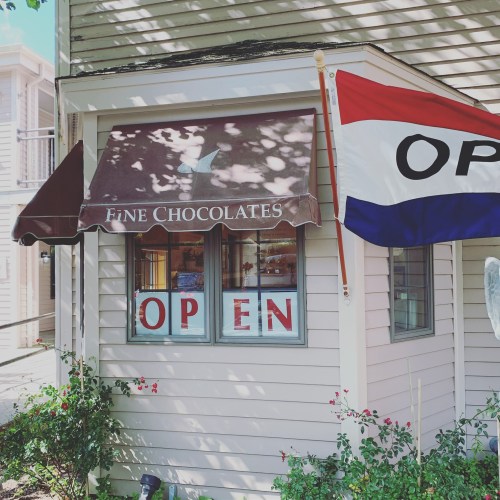 A charming chocolate shop with a brown awning and an "OPEN" sign in the window, surrounded by greenery.