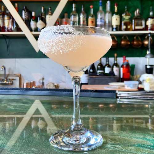 A cocktail in a glass with a sugared rim, set against a bar with various bottles in the background.