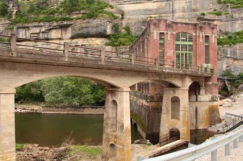Historic brick building beside a river, with a stone arch bridge and lush greenery in the background.
