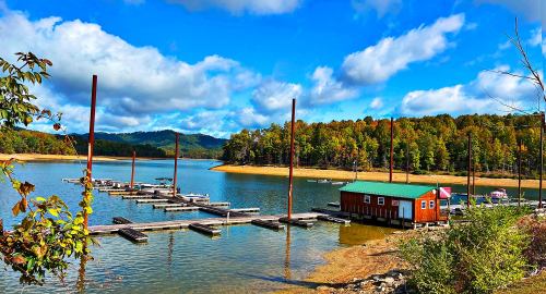 A serene lake scene with a red boathouse, docks, and colorful autumn trees under a blue sky with fluffy clouds.