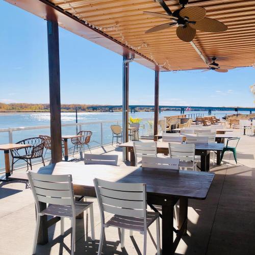Outdoor dining area with wooden tables and chairs overlooking a river, under a sunny blue sky.