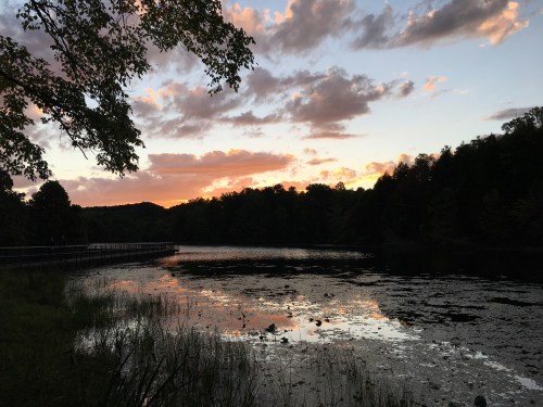 A serene lake at sunset, with colorful clouds reflecting on the water and surrounded by trees and hills.