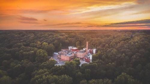 Aerial view of a red brick building surrounded by lush green trees at sunset, with a colorful sky in the background.