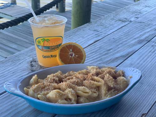 A bowl of pasta topped with breadcrumbs, next to a drink with an orange slice, on a wooden table by the water.