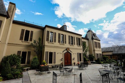A charming building with a large wooden door, surrounded by greenery and outdoor seating under a blue sky.