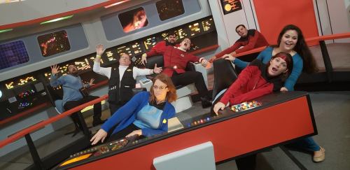 A group of people in Star Trek costumes poses on a spaceship bridge set, interacting playfully with controls.