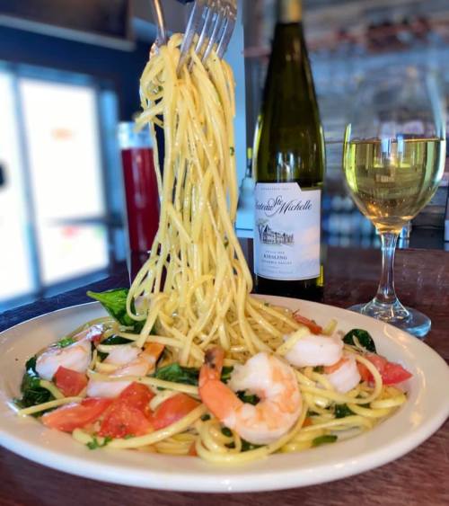 A plate of shrimp pasta with tomatoes and spinach, accompanied by a glass of white wine and a bottle in the background.
