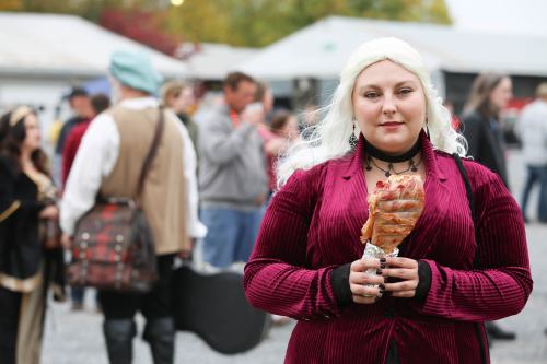 A woman with long blonde hair in a burgundy outfit holds a turkey leg, surrounded by a crowd in a festive setting.