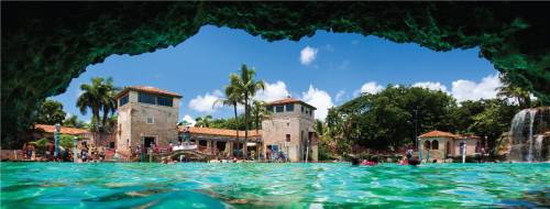 View from a cave opening overlooking a vibrant pool area with palm trees and buildings under a bright blue sky.