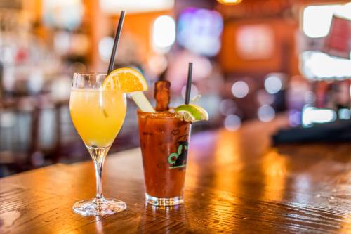 Two cocktails on a wooden bar: a yellow drink with an orange slice and a Bloody Mary with a lime and bacon garnish.
