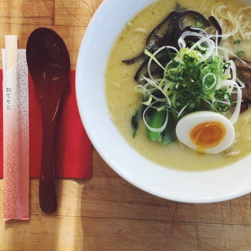 A bowl of ramen with green onions, mushrooms, and a soft-boiled egg, accompanied by chopsticks and a spoon on a wooden surface.