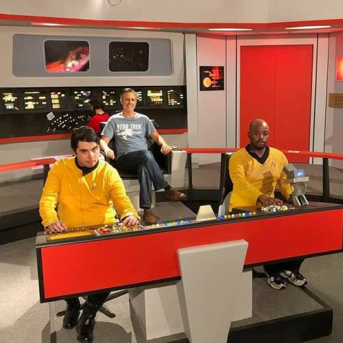 Three people in Star Trek uniforms sit at a control panel, with one person in a casual shirt seated behind them.