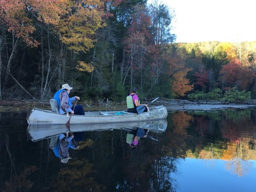 Two people in a canoe on a calm lake, surrounded by autumn trees reflecting in the water.