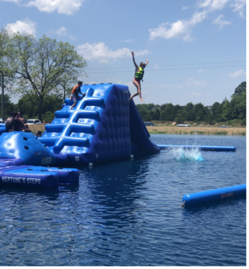 A person jumps off a blue inflatable water slide into a lake on a sunny day, with others nearby enjoying the water.