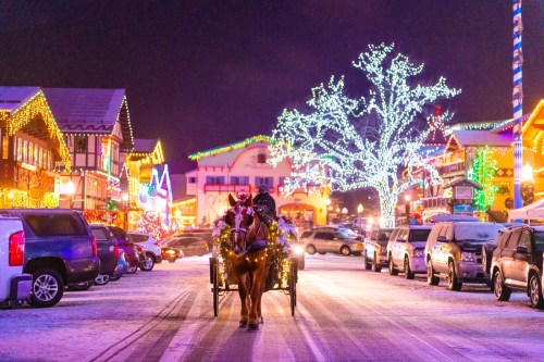 A horse-drawn carriage travels down a snowy street lined with brightly lit buildings and festive decorations.