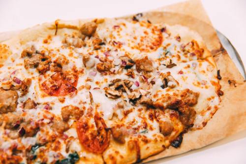 A close-up of a rectangular pizza topped with melted cheese, sausage, tomatoes, and herbs on a parchment-lined tray.