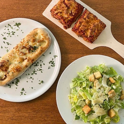A plate with a cheesy flatbread, a Caesar salad with croutons, and two pieces of baked lasagna on a wooden board.