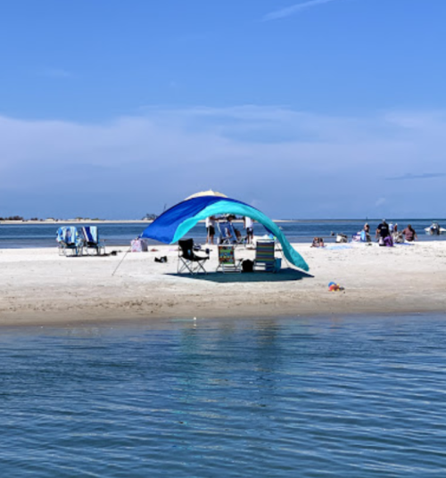 A beach scene featuring a blue and teal tent, chairs, and people relaxing on the sand by the water.