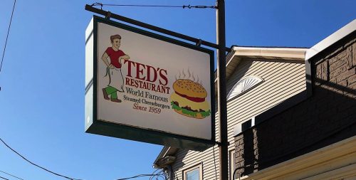 Sign for Ted's Restaurant, featuring a burger, with outdoor seating and a menu board visible. Trees in the background.