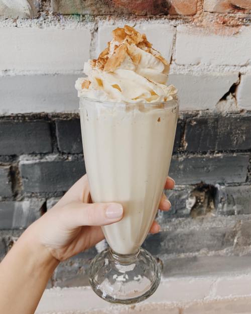 A hand holding a tall glass of creamy milkshake topped with whipped cream and crushed cookies against a brick wall.