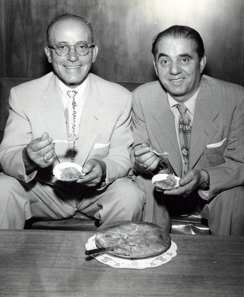 Two men in suits sitting together, smiling and holding plates with pie, with a pie on the table in front of them.