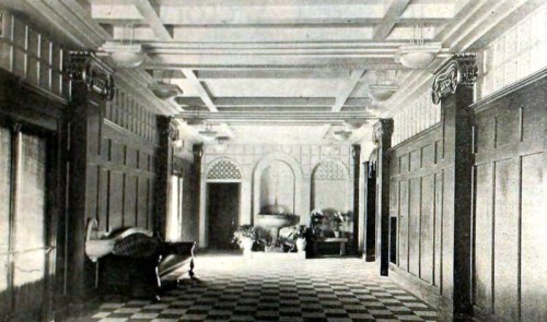 A vintage hallway with checkered flooring, ornate ceiling, and decorative wood paneling, featuring a sofa and plants.