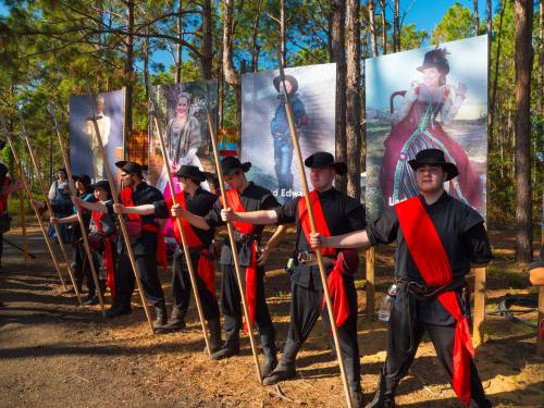 A group of people in black attire with red sashes hold spears, standing in front of large portraits in a forested area.