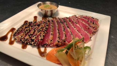 Sliced sesame-crusted tuna on a plate, served with dipping sauce and garnished with vegetables.