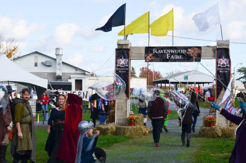 A lively scene at Ravenwood Faire, featuring colorful flags, attendees in costumes, and large bubbles in the air.