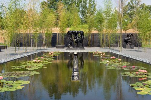 A serene garden with a pond filled with lily pads, flanked by sculptures and surrounded by bamboo and greenery.