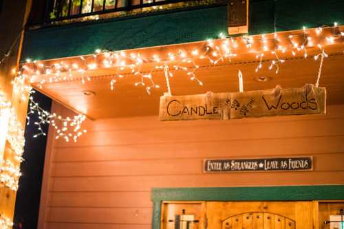 A wooden sign reading "Candle Woods" hangs above twinkling fairy lights on a cozy cabin entrance.
